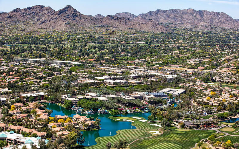 Aerial view of a suburban area with houses, roads, and a golf course surrounded by mountains.